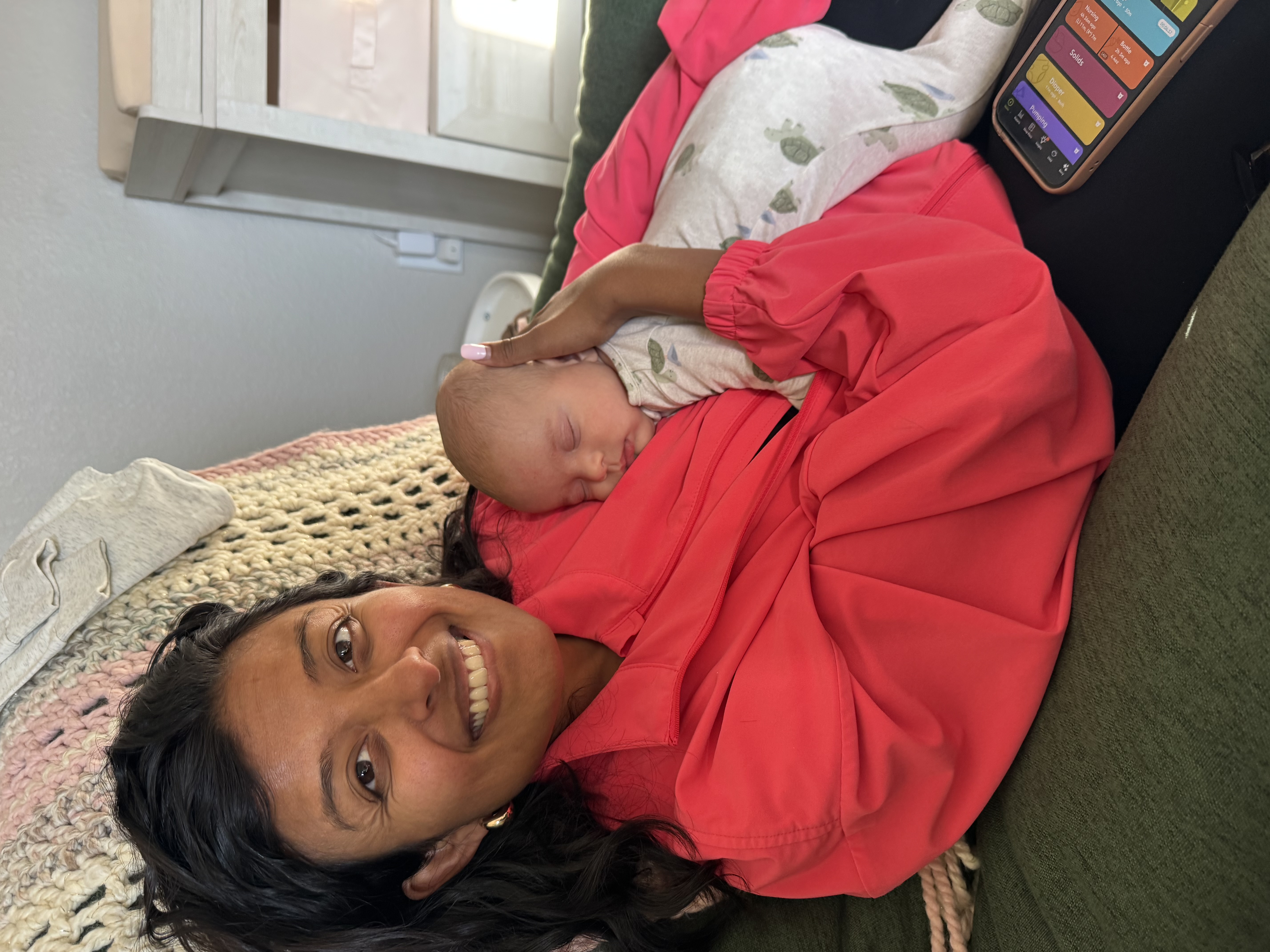 A mother in a pink jacket holding her sleeping baby close, smiling at the camera